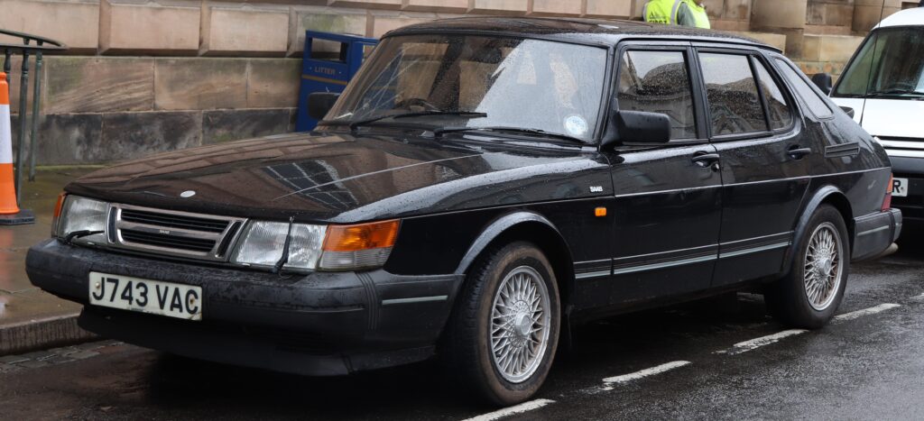 1992 Saab 900 S Turbo 2.0 w kolorze czarnym, zaparkowany na ulicy w deszczowy dzień, widok z przodu i z boku – klasyczny szwedzki sedan z charakterystyczną linią nadwozia i felgami typu siatkowego.