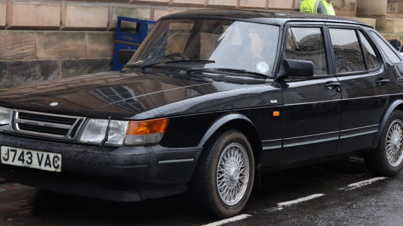 1992 Saab 900 S Turbo 2.0 w kolorze czarnym, zaparkowany na ulicy w deszczowy dzień, widok z przodu i z boku – klasyczny szwedzki sedan z charakterystyczną linią nadwozia i felgami typu siatkowego.