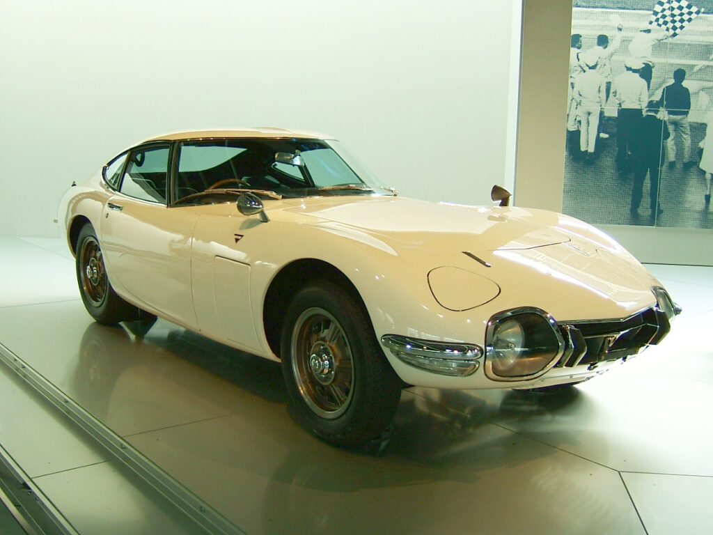 Biała Toyota 2000GT – klasyczne japońskie coupé z lat 60., prezentowana w nowoczesnym muzeum motoryzacyjnym.
