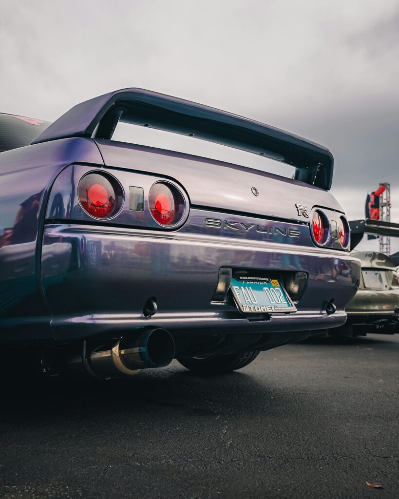 Tył Nissana Skyline GT-R R32 w kolorze Midnight Purple, z widocznym emblematem GT-R, podwójnymi okrągłymi światłami tylnymi i sportowym wydechem, zaparkowany na czarnym asfalcie podczas zlotu samochodowego.