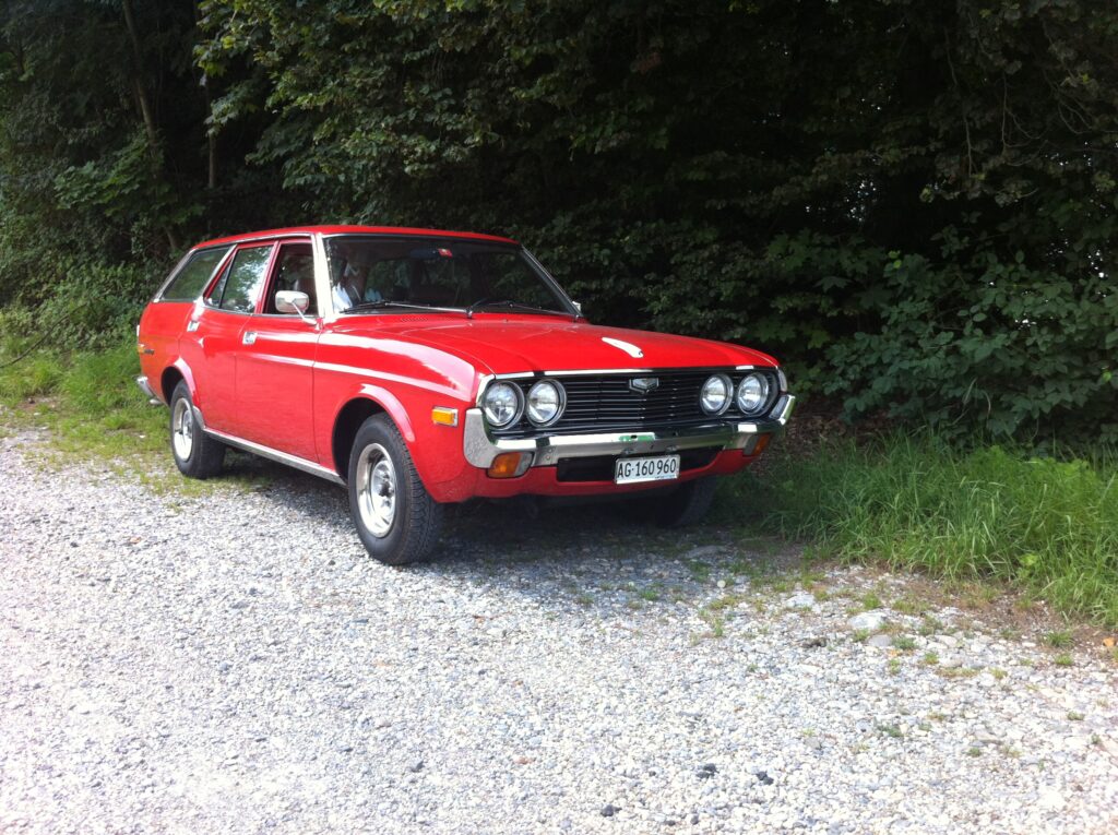 Czerwona Mazda 929 pierwszej generacji w wersji kombi z 1975 roku, zaparkowana na żwirowym podłożu przy lesie